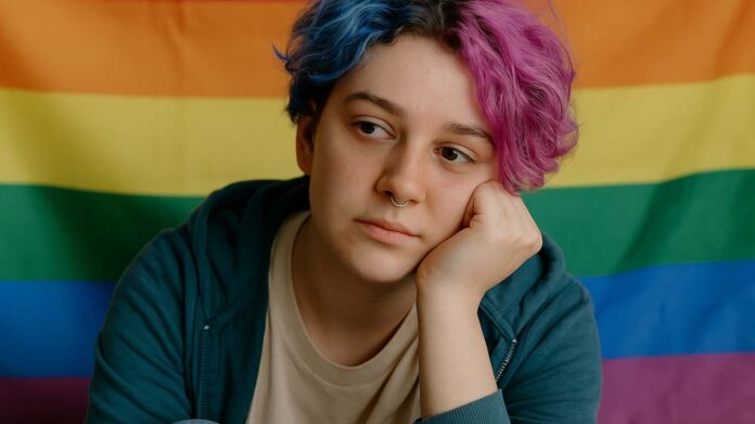 Osoba LGBTQIA+ z tęczowymi włosami na tle flagi tęczowej, zamyślona i refleksyjna, symbolizująca temat psychodelików i zdrowia psychicznego.